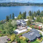 An aerial view of waterfront homes nestled among lush green trees, with a large blue lake in the background and a distant city skyline on the horizon.
