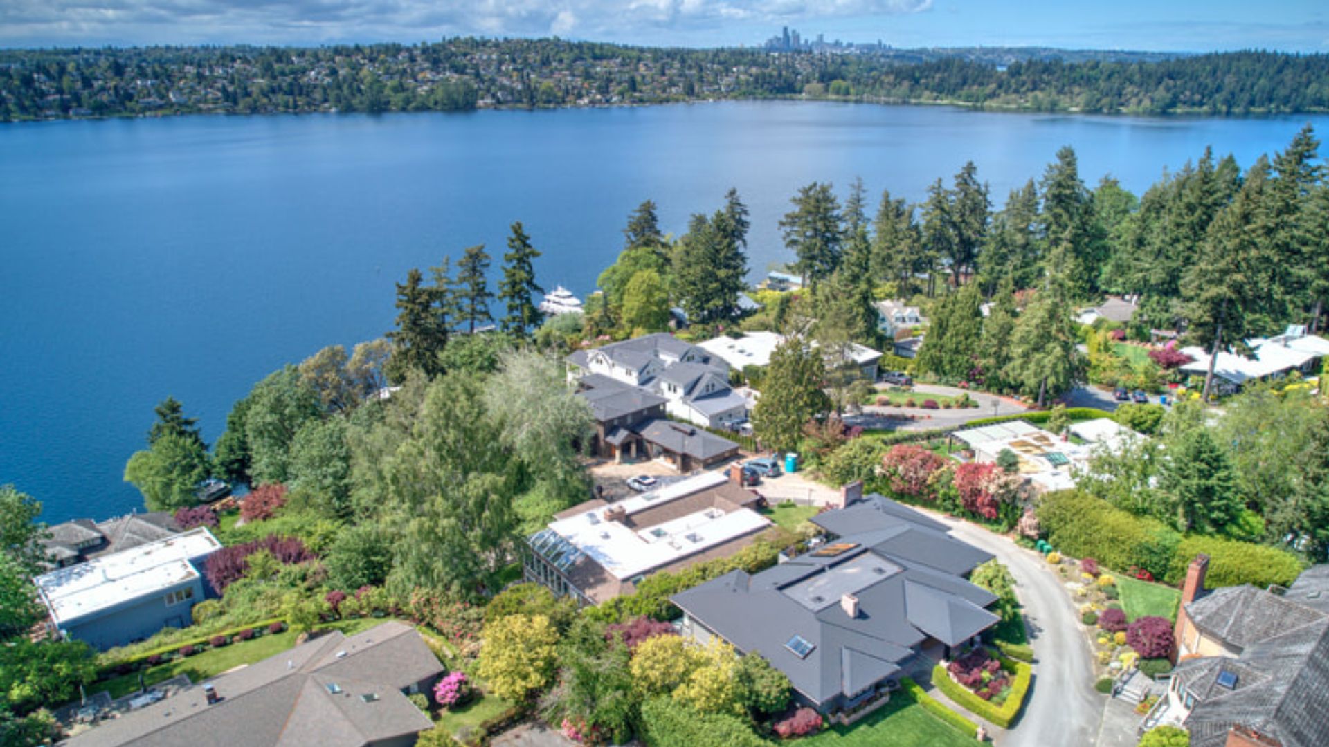 An aerial view of waterfront homes nestled among lush green trees, with a large blue lake in the background and a distant city skyline on the horizon.